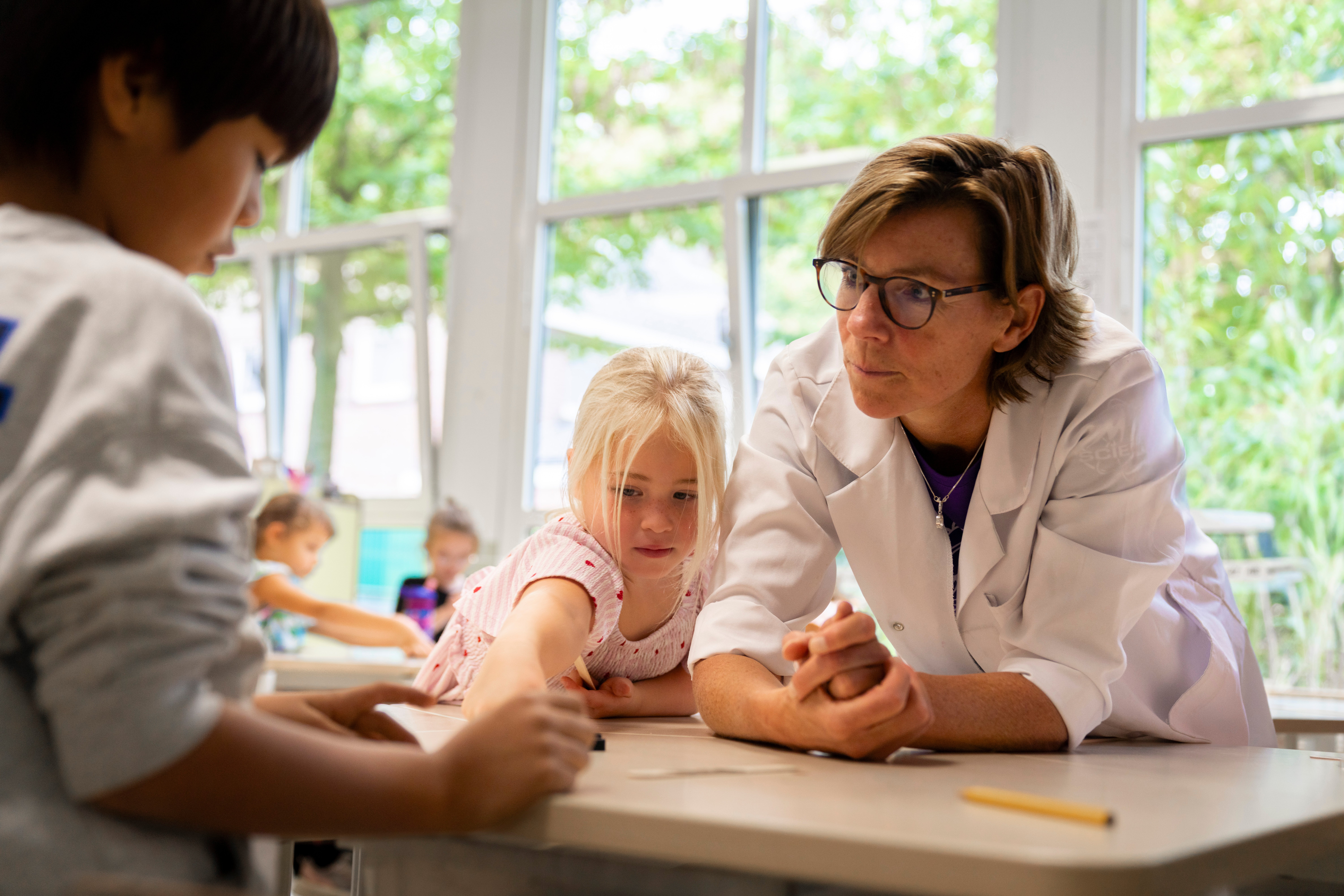 Begeleider van ASML Junior Academy kijkt mee met kinderen tijdens een klaslokaalexperiment. Begeleider van ASML Junior Academy kijkt mee met kinderen tijdens een klaslokaalexperiment.