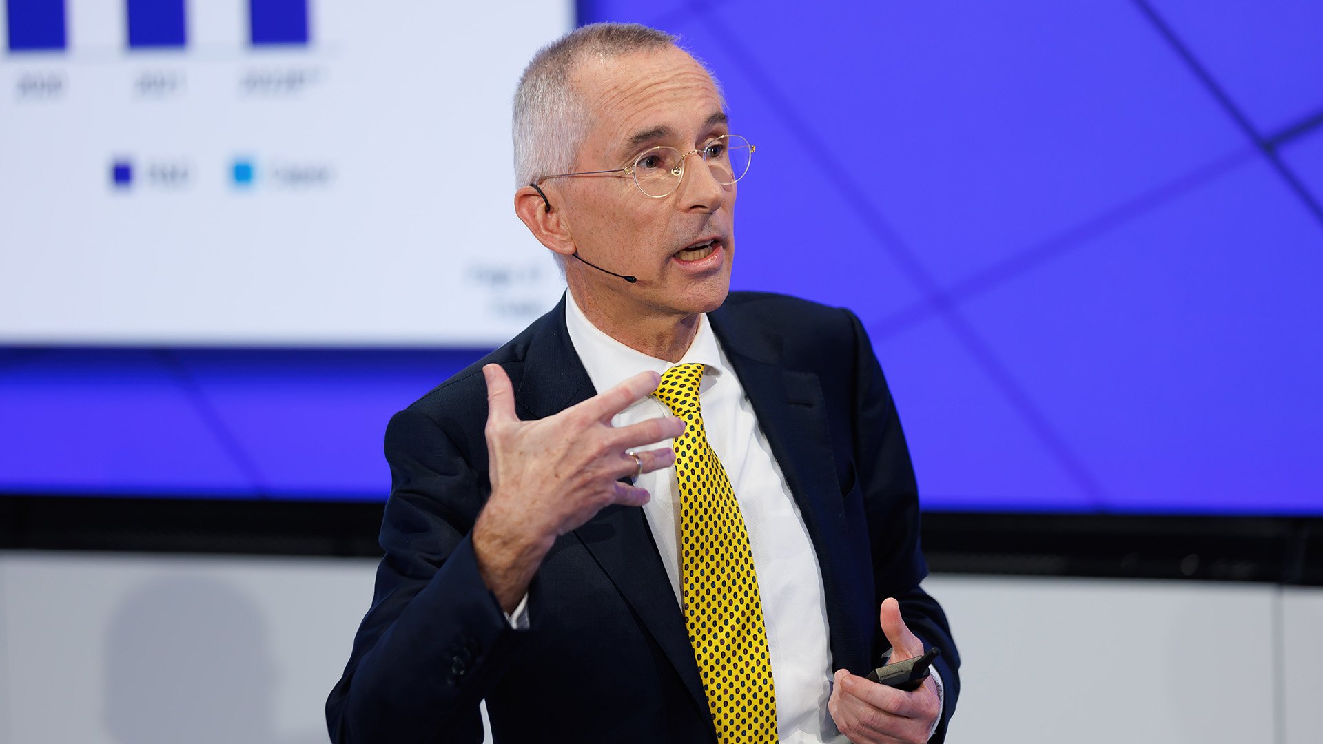 Man in a yellow tie gesturing while speaking on stage Man in a yellow tie gesturing while speaking on stage